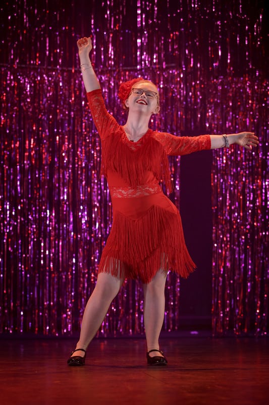 A girl in a red tango dress in dance pose in front of a pink background.